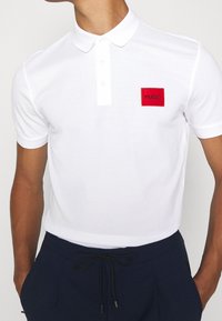 Polo blanc en coton, avec un patch rectangulaire rouge comportant le texte "HUGO". Manches courtes et col classique.