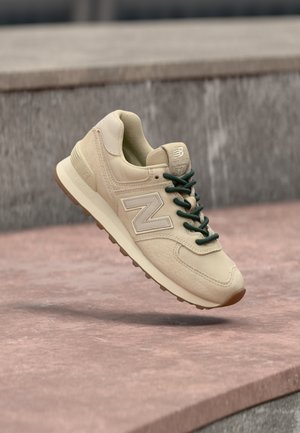 Μπεζ αθλητικό παπούτσι New Balance με σκουροπράσινα κορδόνια, επάνω μέρος από σουέντ και δίχτυ, απορρόφηση κραδασμών στη μέση σόλα, και καφετί εξωτερική σόλα σε πέτρινη επιφάνεια.