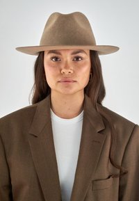 Brown felt hat with wide brim, worn with a brown blazer over a white ribbed shirt. The model has long, wavy hair and gold earrings.