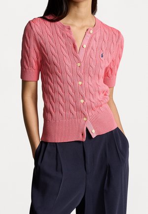 Frau trägt eine rosa Kurzarm-Strickjacke mit Knöpfen, kombiniert mit hoch taillierter dunkelblauer Hose, Hände in den Taschen.