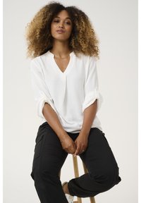 Blouse blanche texturée à col en V avec des manches retroussées, associée à un pantalon cargo noir et des baskets blanches, assise sur un tabouret en bois.