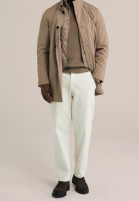 Béžový kabát s černými knoflíky přes hnědý svetr, doplněný bílými kalhotami a tmavě hnědými botami. Hladká textura, na míru šitý design.