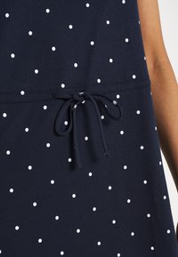 Robe en tissu bleu marine à pois blancs, avec une ceinture à cordon de serrage à la taille et une texture lisse.