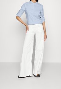 Kvinna som bär en ljusblå långärmad skjorta och högmidjade vita vida jeans med fransade nederkanter, stående med handen på höften.