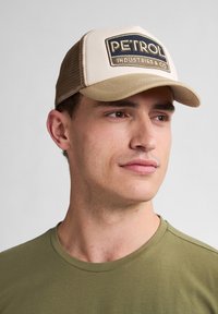 Gorra trucker beige con visera curva, que presenta un parche rectangular negro con la inscripción "PETROL INDUSTRIES & CO" en letras blancas.