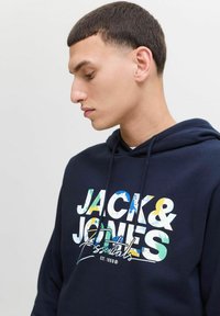 Marineblaue Kapuzenjacke mit einem mehrfarbigen Drucklogo, das "JACK & JONES" und "Essentials" Text enthält. Das Material sieht texturiert und warm aus.