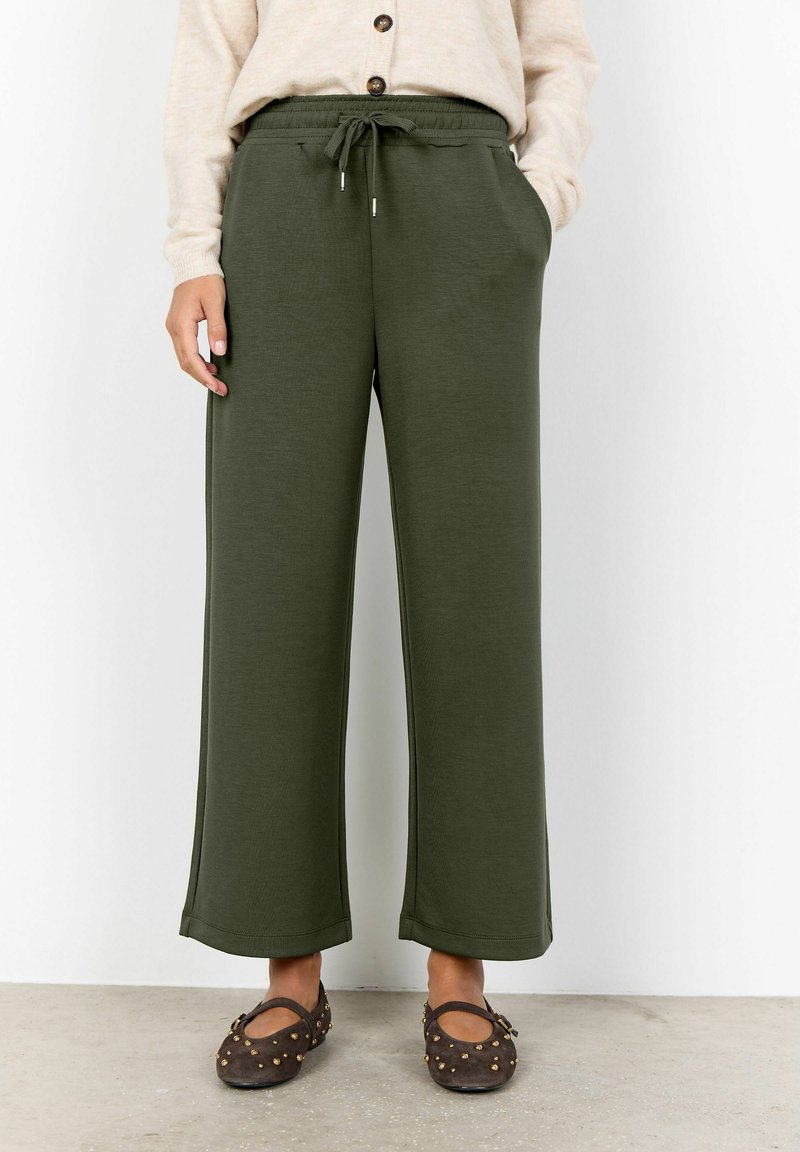 Personne portant un pantalon large vert olive avec une taille à cordon, un pull beige boutonné et des chaussures Mary Jane cloutées marron foncé.