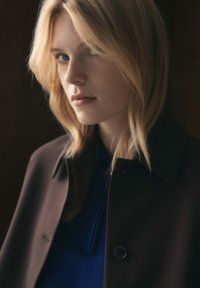 Een close-up van een persoon in een donkerbruine jas met een blauw shirt. Heeft lichtblond haar en een neutrale gezichtsuitdrukking. Zacht, natuurlijk licht.