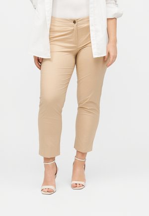 Naine, kes kannab beežikaid kitsaid lühikesi pükse, valgeid rihmikuid ja valget särki valge topi kohal, seisab tavalise tausta ees.