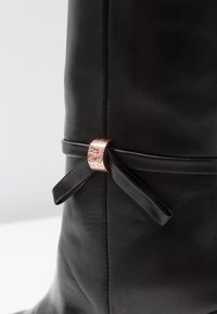 Black leather knee-high boot featuring a metallic rose gold accent and a tied ribbon detail on the side. Smooth texture with stitching.