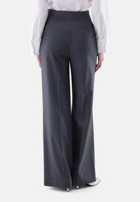 Person, die hoch taillierte, weit geschnittene, dunkelgraue Hosen trägt, kombiniert mit einem weißen Langarmhemd, Rückansicht.