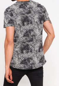 Hombre con una camiseta de manga corta negra y blanca con manchas y pantalones negros, mostrado desde atrás con un tatuaje en la parte superior del brazo derecho.
