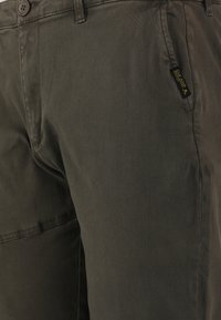 Olivgröna byxor i bomullsblandning, med framknäppning, sidoficka och en liten etikettdetalj. Mjuk, texturerad yta.