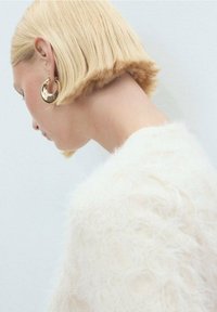 Des boucles d'oreilles créoles en or pendent des oreilles, complétant un pull texturé en laine pelucheuse de couleur blanc cassé. Les cheveux sont coiffés en un carré court et droit.