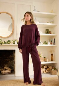 Burgundyrødt loungewear-sæt lavet af ribbet stof, der har en afslappet fit top med snoretræk i taljen og brede bukser med en glat tekstur.