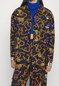 Svart jacka med guldkedja och djurmönster, prydd med färgglada blommor. Inkluderar adidas-logotyp på fickan och randiga detaljer vid ärmslutet.