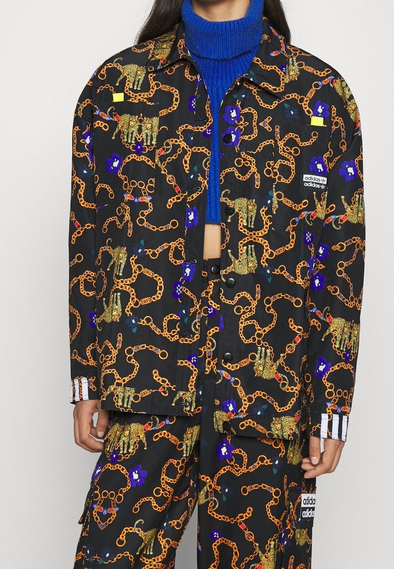 Svart jacka med guldkedja och djurmönster, prydd med färgglada blommor. Inkluderar adidas-logotyp på fickan och randiga detaljer vid ärmslutet.