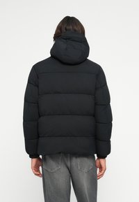 Persona che indossa un giubbotto imbottito nero con cappuccio e jeans grigi, vista di spalle su uno sfondo bianco semplice.