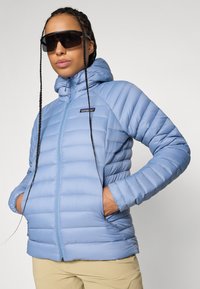 Osoba nosi svijetloplavu jaknu s kapuljačom i bež hlače, ruke u džepovima, i velike crne sunčane naočale.