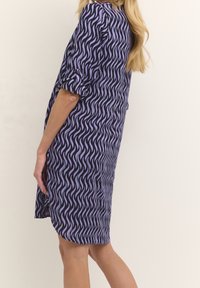 Lila Kleid mit einem wellenförmigen Muster in helleren Lila-Tönen. Weicher Stoff, hochrollbare Ärmel und seitliche Schlitz für mehr Komfort. Knielanges Design.