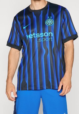 Mann trägt ein schwarz-blau gestreiftes Fußballtrikot mit Nike-Logo, Sponsortext und blauen Shorts.