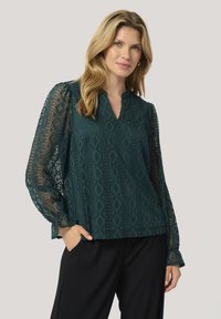 Blusa in pizzo color teal con maniche decorate, scollatura a V e vestibilità rilassata, caratterizzata da un motivo a diamante e una leggera lucentezza. Abbinata a pantaloni neri.