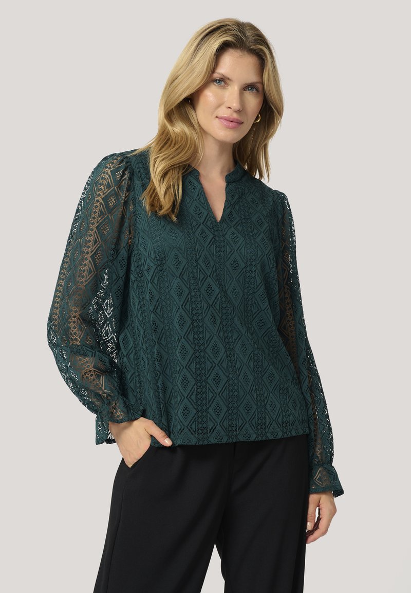 Blusa in pizzo color teal con maniche decorate, scollatura a V e vestibilità rilassata, caratterizzata da un motivo a diamante e una leggera lucentezza. Abbinata a pantaloni neri.