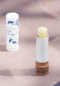 Lippenbalsam mit einem blassgelben Stift, untergebracht in einem weißen Behälter mit blauen botanischen Designs und einem hellbraunen Boden, der mit "Kakaobutter" beschriftet ist.