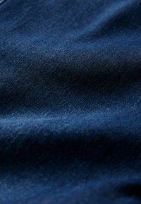 Tmavě modrá denimová látka s hladkou texturou, ukazující jemné barevné variace a mírný lesk. Detailní pohled odhaluje podrobný vzor tkaní.