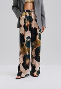 Leveälahkeiset housut, jotka on tehty sileästä kankaasta ja joissa on suuri kukkakuosi vihreän, mustan ja beigen sävyissä, yhdistetty harmaaseen bleiseriin.