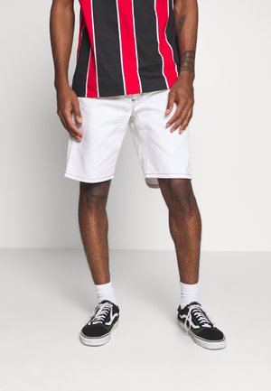 Homme portant un short blanc mi-cuisses, une chemise à rayures verticales noires et rouges, des chaussettes blanches et des baskets noires, debout devant un fond uni.