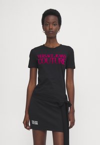 Svart bomull t-skjorte med rund hals og korte ermer. Har fet fuchsia tekst "VERSACE JEANS COUTURE" på forsiden. Tilbehør er et knyttet svart skjørt.