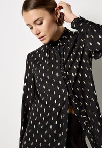 Black blouse with gold leaf pattern, ruffled neckline, and cuffed sleeves. Features button closure and a relaxed fit design.