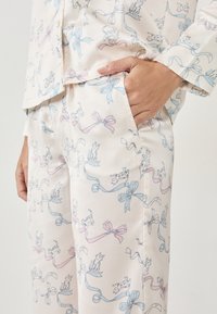 Conjunto de pijama en tela color crema con un patrón de cintas azules y rosas y pequeñas ilustraciones de perros. Textura suave, corte relajado y bolsillos.