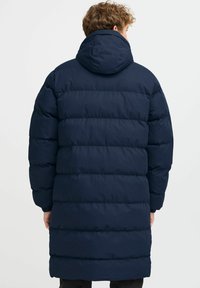 Tengeréskék pufi kabát, kapucnival, vízszintes steppelt mintával és elasztikus mandzsettákkal. Az anyag sima és vízlepergető hatású.
