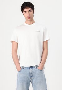 White cotton t-shirt with short sleeves, round neck, and small "TOMMY JEANS" logo on the chest. Worn with light blue, relaxed-fit jeans.