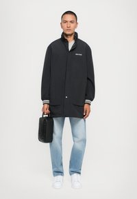 Črn lahkoten dežni plašč z gumbi in stranskimi žepe, v kombinaciji s svetlo modrimi kavbojkami in belimi športnimi copati. Drži majhno črno torbico.