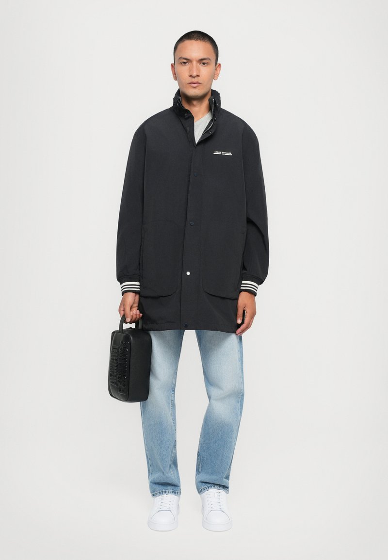 Črn lahkoten dežni plašč z gumbi in stranskimi žepe, v kombinaciji s svetlo modrimi kavbojkami in belimi športnimi copati. Drži majhno črno torbico.