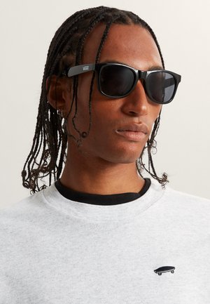 Schwarze Sonnenbrille mit dunklen Gläsern und Vans-Logo am Bügel. Getragen mit einem hellgrauen Sweatshirt, das ein kleines besticktes Skateboard-Detail aufweist.