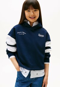 Granatowy hoodie z białymi akcentami i haftowanymi logo; nałożony na jasnoniebieską koszulę z małymi czarnymi wzorami serc; dżinsy.