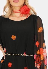 Czarna sukienka w kwiatowe wzory w odcieniach pomarańczowym i różowym, z przezroczystymi długimi rękawami i cienkim złotym pasem. Czerwony kwiatowy akcent przy dekolcie.