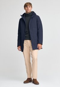Cappotto imbottito blu navy con cappuccio, con chiusura zip, indossato sopra un dolcevita scuro. I pantaloni crema e gli stivaletti in suede marrone completano il look.