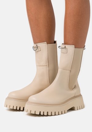 Botines beige de cuero, con suela gruesa de borde dentado, laterales elásticos y detalles metálicos cerca de la parte superior.