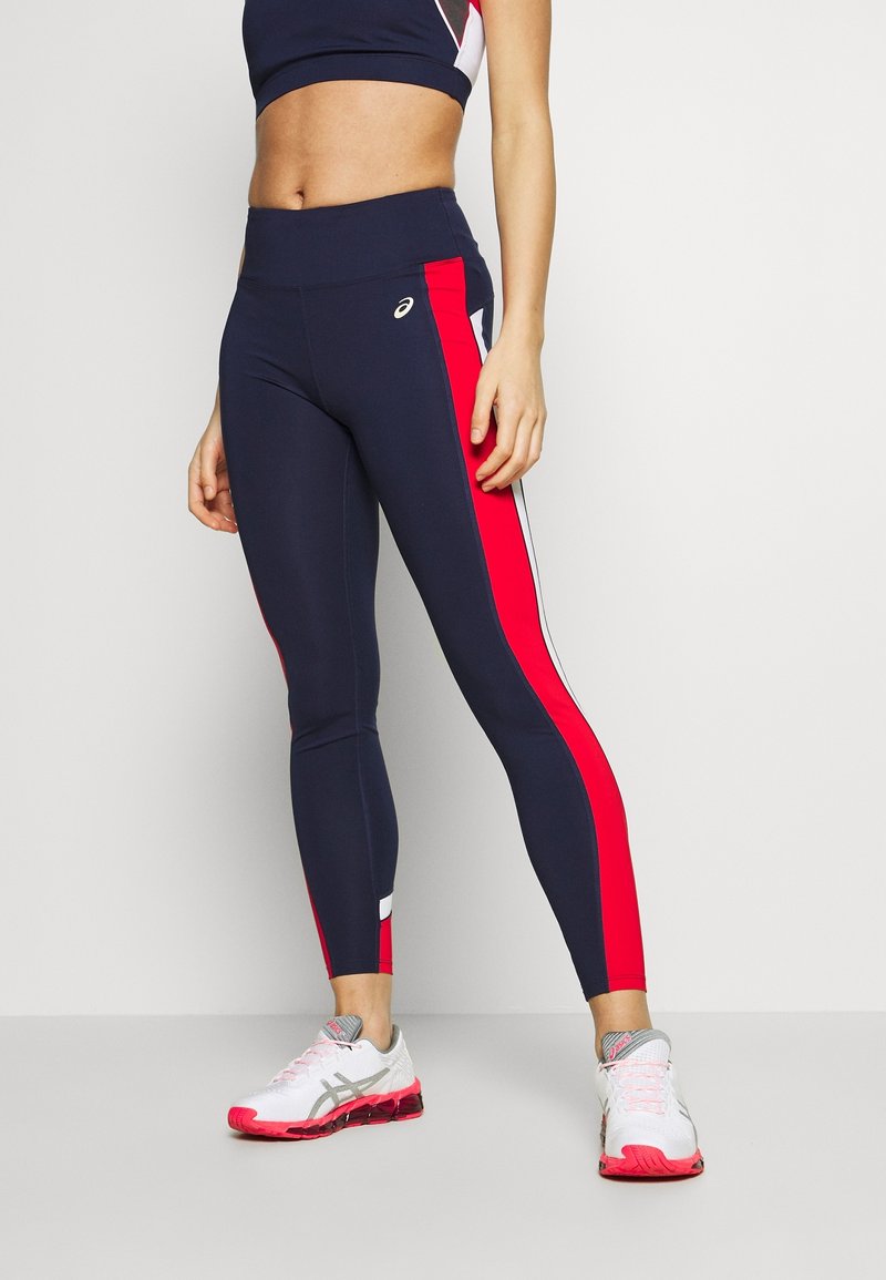 Kvinde iført marineblå leggings med røde og hvide sidebaner og hvide løbesko med pink såler, stående foran en ensfarvet baggrund.