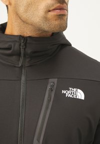 Giacca nera con zip e colletto alto, dotata di una piccola tasca con zip e un logo bianco sul petto. Tessuto liscio e elasticizzato.