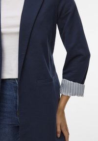 Mornarsko moder plašč z ovratnikom in enim žepom, s pripetimi rokavi v svetlo modri in beli črti. Gladka tekstura blaga.