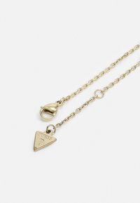 Collier en chaîne en or avec un pendentif triangulaire portant une gravure de marque. La chaîne possède un fermoir à pince et une texture lisse.