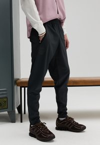 Zwarte ademende joggers met een elastische tailleband, zakken met rits en een logodetail, gecombineerd met bruine sneakers en een lichtroze top.