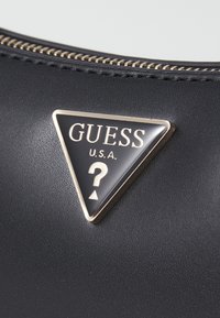 Material negro texturizado con un logo negro acentuado en dorado en forma triangular. El logo presenta "GUESS" y un diseño de signo de interrogación.
