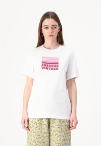 Balta medvilninė marškinė su rožiniu grafiniu užrašu "AMERICAN VINTAGE" ir horizontaliomis juostelėmis viršuje. Modelis dėvi raštuotas pilkas kelnes.